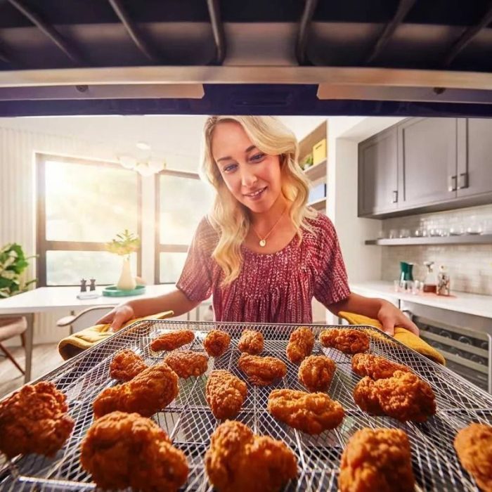 Mujer sacando bandeja de pollo frito dorado desde un horno eléctrico en una cocina moderna